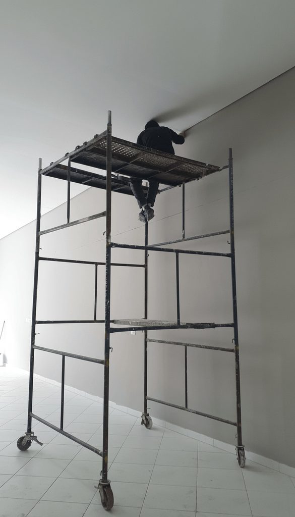 Profissional sobre andaime realizando acabamento e pintura interna de ambiente comercial em São Paulo